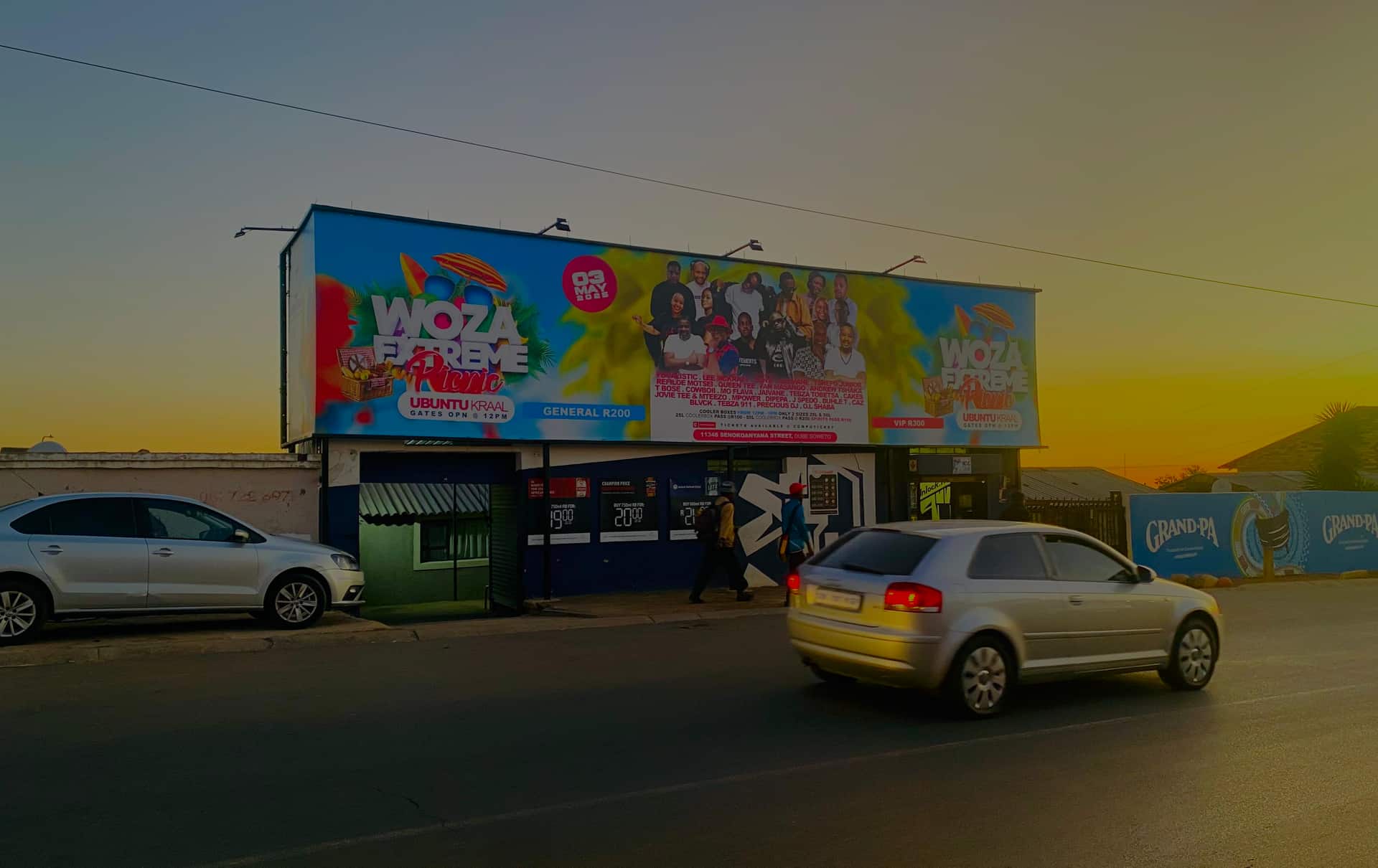 Woza Extreme Picnic billboard on a Gauteng shop roof at golden hour, a silver car passing below